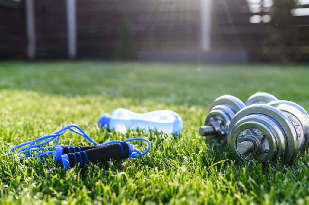 Fitness-Equipment im Garten, darunter eine Hantel, ein Springseil und eine Wasserflasche auf grünem Rasen – ideal für ein Training unter einem Pavillon.